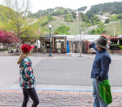 A man and women standing on sidewalk looking across at Aspen Mountain Ski area with some snow on the ski runs.